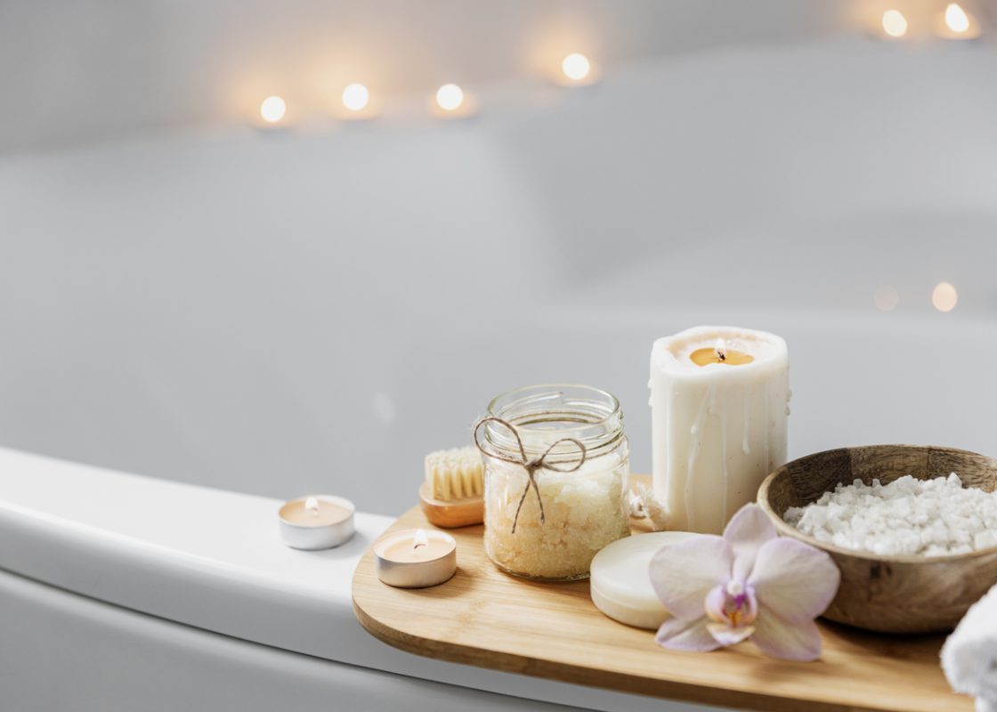 Preparation for hotel spa treatment or home bath procedure. White washbasin in bathroom with accessories on tray. Burning candles, washcloth, soap, foot brush, bottle with sea salt, orchid flower